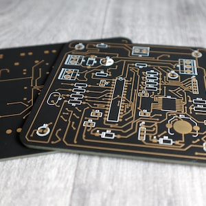 以下が含まれることがあります： 金色のトレースと白いマークが付いた黒い回路基板が2枚。 上の基板には、R1、R2、C1、C2、C3など、数字と文字でラベル付けされたコンポーネントがあります。 下の基板は、よりシンプルなデザインです。