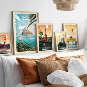Pode incluir: Cinco cartazes de viagem emoldurados com ilustra&ccedil;&otilde;es de marcos famosos. Os cartazes est&atilde;o dispostos em uma parede branca acima de uma cama com almofadas marrons e brancas. Os cartazes representam a Golden Gate Bridge, uma pagoda no Vietn&atilde;, o Port&atilde;o de Brandemburgo, a Table Mountain na &Aacute;frica do Sul e uma montanha em Ibiza.