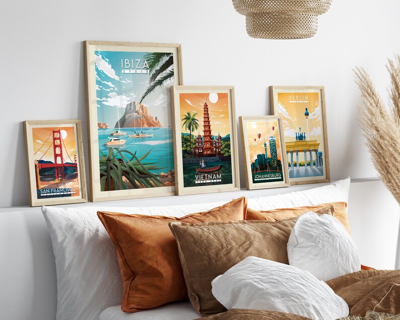Pode incluir: Cinco cartazes de viagem emoldurados com ilustra&ccedil;&otilde;es de marcos famosos. Os cartazes est&atilde;o dispostos em uma parede branca acima de uma cama com almofadas marrons e brancas. Os cartazes representam a Golden Gate Bridge, uma pagoda no Vietn&atilde;, o Port&atilde;o de Brandemburgo, a Table Mountain na &Aacute;frica do Sul e uma montanha em Ibiza.