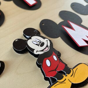 May include: Black and white Mickey Mouse character cutout with red shorts and yellow shoes. The cutout is decorated with black glitter.