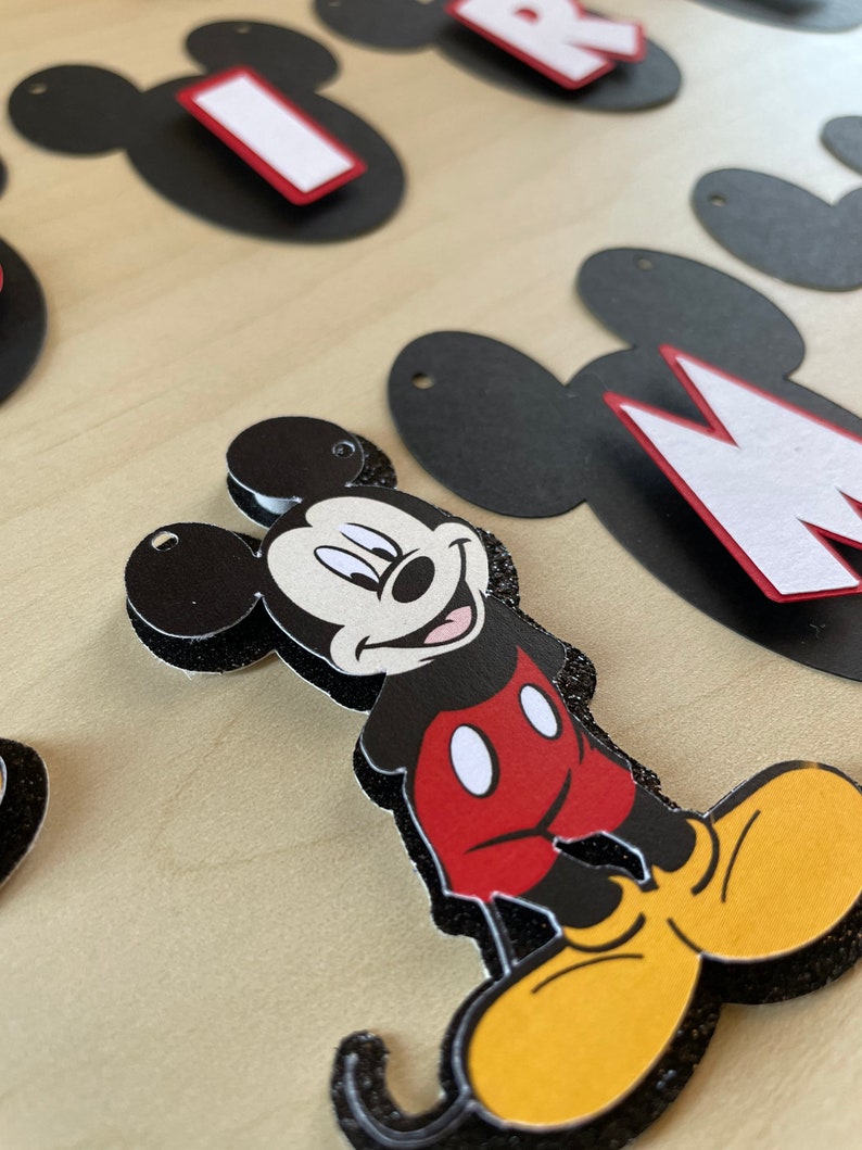 May include: Black and white Mickey Mouse character cutout with red shorts and yellow shoes. The cutout is decorated with black glitter.