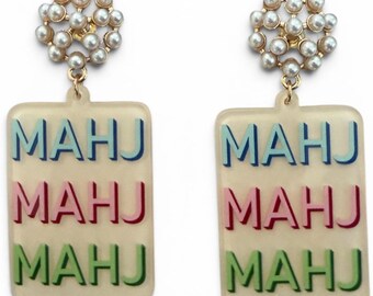 May include: A pair of cream-colored earrings with the word "MAHJ" in blue, pink, and green. The earrings have a rectangular shape and are topped with a cluster of small, round pearls on a gold-colored setting.
