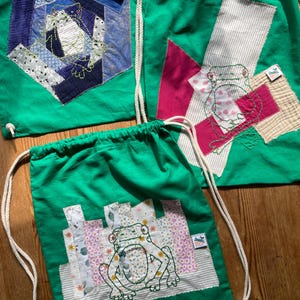 Könnte beinhalten: Drei grüne Kordelzugbeutel mit Froschapplikationen. Jeder Beutel hat ein anderes Patchwork-Design in Blau-, Rosa-, Weiß- und Beigetönen. Die Frösche sind mit schwarzem Faden bestickt. Die Beutel haben weiße Kordelzüge.