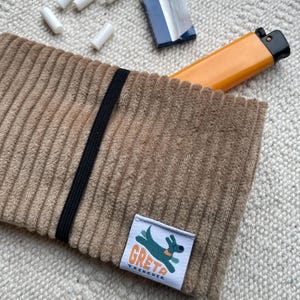 Könnte beinhalten: Eine beige Kordel-Tasche mit einem schwarzen Gummiband, einem Feuerzeug und einer Packung weißer Filter. Die Tasche hat ein Etikett mit einer grünen Hunde-Grafik und dem Text "GRETA TREKKER".