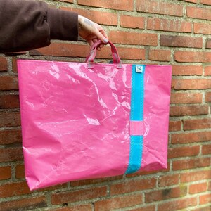 Könnte beinhalten: Eine wiederverwendbare rosa Tragetasche mit einem blauen Streifen an der Seite. Die Tasche hat einen Griff und ist aus wasserfestem Material.