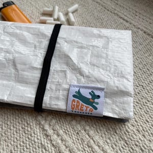Könnte beinhalten: Eine weiße, strukturierte Tasche mit einem schwarzen Gummiband. Die Tasche hat ein Etikett mit einer grünen Hunde-Grafik und dem Text "GRETA TREKKER". Ein orangefarbenes Feuerzeug und mehrere weiße Filter sind sichtbar.