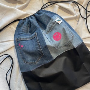 Könnte beinhalten: Eine schwarze und blaue Kordeltasche aus Jeans mit einem rosafarbenen Smiley und zwei rosafarbenen Herzen. Die Tasche hat einen schwarzen Kordelzug und ein schwarzes Stofffutter.