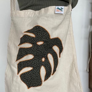 Könnte beinhalten: Eine beige Canvas-Tragetasche mit einem schwarzen und grünen Leopardenmuster-Monstera-Blatt-Design. Die Tasche hat einen orangefarbenen Umriss und ein kleines grünes Etikett mit dem Text "Greta's Treasures" auf der Vorderseite.