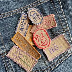 Könnte beinhalten: Nahaufnahme einer blauen Jeans-Tasche, verziert mit mehreren gestickten Aufnähern. Die Aufnäher zeigen verschiedene Designs, darunter eine Qualle, eine Blume und Texte wie "Karma" und "No.". Die Aufnäher sind in Beige, Blau, Orange, Rot und Lila gehalten.