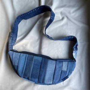 Könnte beinhalten: Eine blaue Jeans-Umhängetasche aus recyceltem Denim. Die Tasche hat einen Reißverschluss und einen langen, verstellbaren Riemen.
