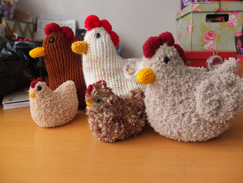 May include: Five crocheted chicken toys in various colors, including brown, white, beige, and brown with red accents. The chickens are all sitting on a wooden surface.