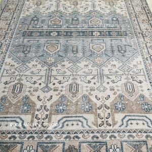 May include: A rectangular area rug with a detailed geometric pattern in shades of beige, blue, and brown. The rug features a central design with repeating shapes and borders, with the text "RUGS VALLEY STUDIO" visible.