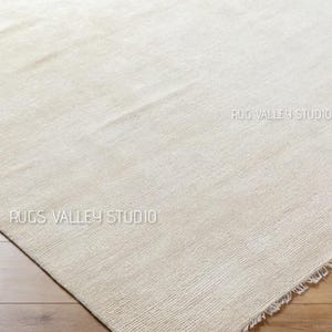 May include: A neutral-toned rug with a textured surface and a fringed edge. The rug is a light beige color and appears to be made of a woven material. The words "RUGS VALLEY STUDIO" are printed on the rug.