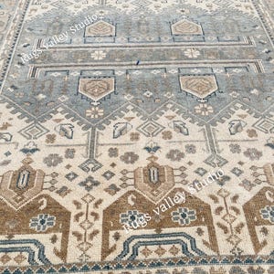 May include: A rectangular area rug with a detailed geometric pattern. The rug features a combination of beige, blue, and brown tones. The design includes repeating shapes and floral motifs, creating a traditional aesthetic. The text "Rugs Valley Studio" is visible.