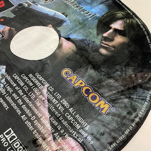 May include: A Resident Evil PlayStation 2 game disc featuring a character holding a gun. The disc has the Capcom logo and the text "Resident Evil" in a stylized font. The disc is black, blue, and white.