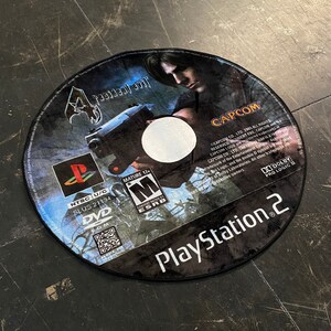 May include: Circular rug featuring the Resident Evil 4 video game disc design. The rug displays the game's title, character art, and PlayStation 2 logo. Includes the ESRB rating and other game details.