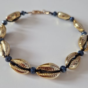 Könnte beinhalten: Eine goldene Muschelkette mit schwarzen Perlen. Die Kette besteht aus Kaurischnecken und schwarzen Perlen.