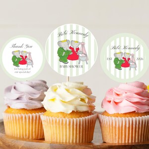 May include: Three cupcakes with pastel frosting, decorated with circular toppers for a baby shower. The toppers feature an elephant couple and text that reads "Thank You", "Bébé Kennedy BABY SHOWER", and "Bébé Kennedy EST 2026".