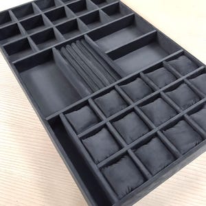 May include: A dark gray jewelry organizer tray featuring multiple compartments for storage. The tray includes a section with small square slots, a row of ring rolls, two rectangular open spaces, and a section containing twelve small square cushions.