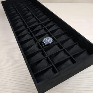 May include: A black watch display tray with multiple compartments, each lined with black fabric. A single wristwatch with a blue face and silver band rests in one compartment. The tray is rectangular and sits on a light-colored wooden surface.