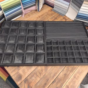 May include: Two-piece black velvet jewelry display tray with multiple compartments. One side features square padded compartments, the other has a mix of compartments and a ribbed section. Fabric swatches are in the background.