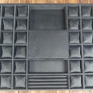 May include: A gray velvet watch display case with multiple compartments. The case has a rectangular shape with individual square compartments for watches, along with larger compartments and a section with horizontal ridges.