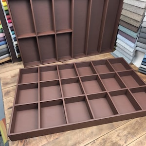May include: Two brown wooden display trays with multiple compartments. One tray has a rectangular shape with 20 compartments, while the other has a larger rectangular shape with fewer compartments. The trays are on a wooden surface.