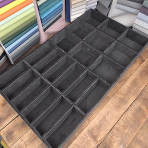 May include: A dark gray wooden display tray with multiple rectangular compartments. The tray is on a wooden surface, with fabric swatches of various colors in the background.
