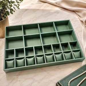 May include: A rectangular sage green jewelry organizer tray featuring multiple compartments of varying sizes, including a row of seven small padded cushions designed for holding watches or bracelets, all finished in a smooth, matte green material.