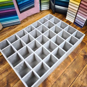 Peut inclure: Un organisateur de rangement rectangulaire gris doté d'une grille de trente-cinq compartiments carrés. L'organisateur est construit avec de fines cloisons créant des espaces ouverts uniformes pour trier des objets, vu en plongée.