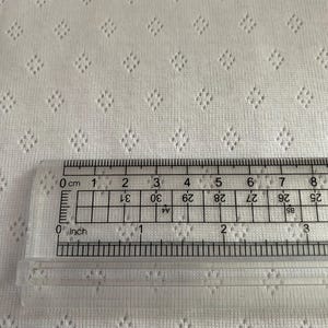 Op de afbeelding: Close-up van witte stof met een ruitvormig oogjespatroon. Een doorzichtige liniaal is op de stof geplaatst, met zowel inches als centimeters. De stof heeft een getextureerd uiterlijk.