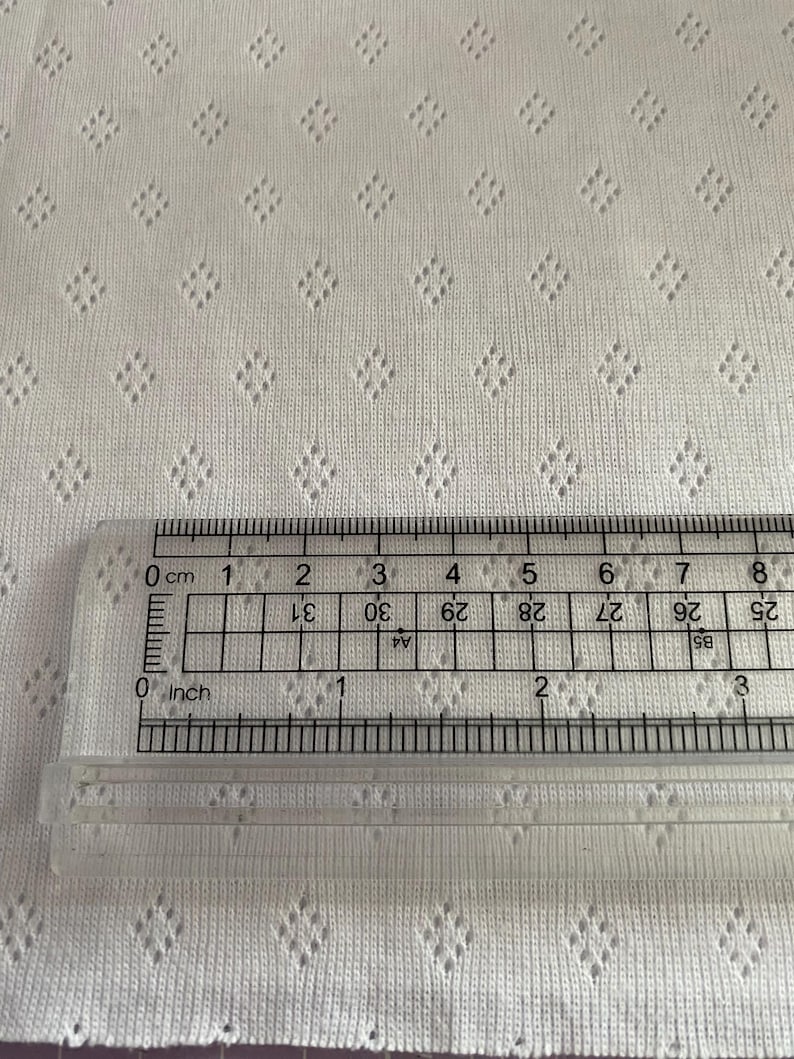 Op de afbeelding: Close-up van witte stof met een ruitvormig oogjespatroon. Een doorzichtige liniaal is op de stof geplaatst, met zowel inches als centimeters. De stof heeft een getextureerd uiterlijk.