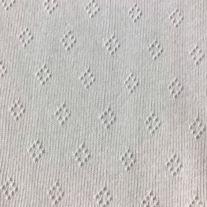 Op de afbeelding: Close-up van een witte gebreide stof met een herhalend ruitpatroon. De stof heeft een getextureerd oppervlak met kleine, gelijkmatig verdeelde ruitvormige openingen. Het gebreide materiaal lijkt zacht en lichtgewicht, geschikt voor kleding of woondecoratie.