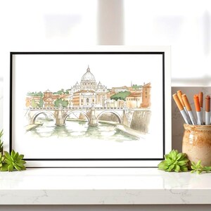 May include: Watercolour print of a cityscape with a bridge and domed building, framed in black and white. The artwork is on a white surface, with a small succulent plant and a ceramic pot with markers.