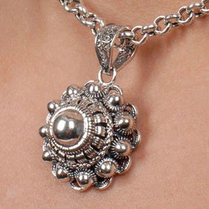 May include: A silver pendant necklace with a round, ornate design. The pendant features a central circle surrounded by intricate details and small beads.