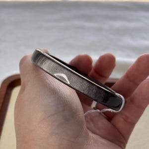 May include: A dark gray bangle bracelet with a rectangular shape. The bracelet has a smooth, polished surface with subtle variations in color. A white string tag is attached to the bracelet.