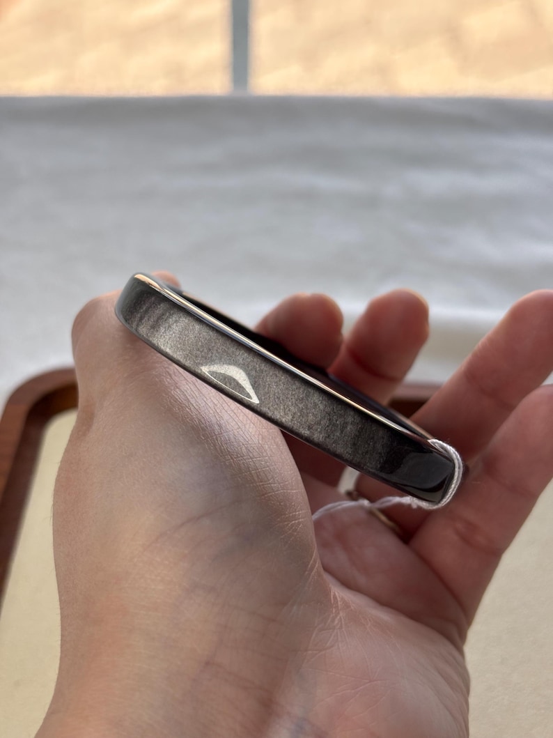 May include: A dark gray bangle bracelet with a rectangular shape. The bracelet has a smooth, polished surface with subtle variations in color. A white string tag is attached to the bracelet.