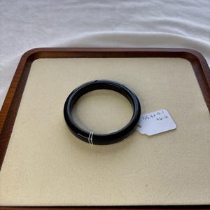 May include: A black, circular bangle bracelet with two thin, parallel white lines. The bracelet is displayed on a beige surface within a wooden tray. A white tag with measurements is attached.