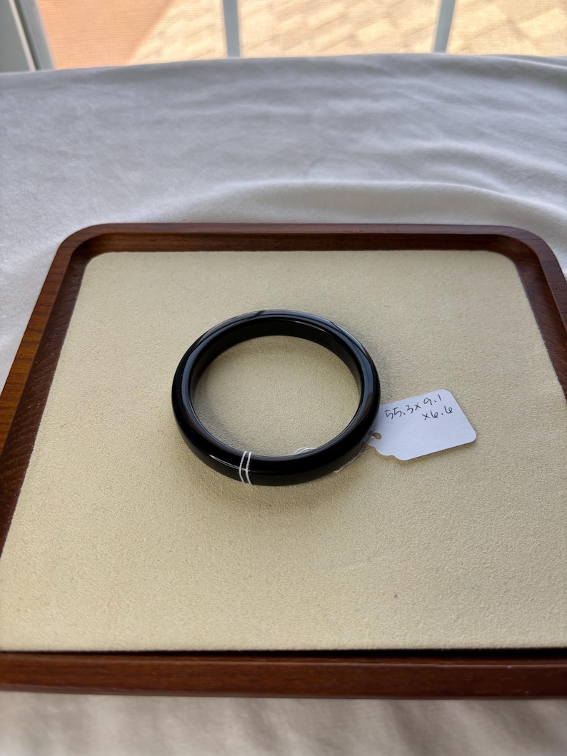 May include: A black, circular bangle bracelet with two thin, parallel white lines. The bracelet is displayed on a beige surface within a wooden tray. A white tag with measurements is attached.