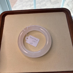 May include: A translucent, pale green jade bangle bracelet. The bracelet is circular and smooth, with a polished finish. A small tag with handwritten measurements is attached. The bangle is displayed on a beige surface within a wooden tray.