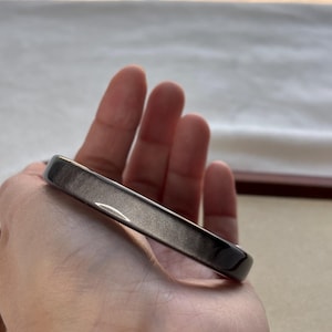 May include: A dark gray, rectangular bangle bracelet with a smooth, polished surface. The bracelet is held in a hand, showcasing its sleek design and metallic sheen. The jewelry piece is a simple, elegant accessory.