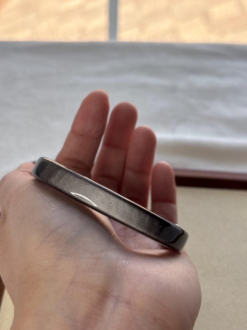 May include: A dark gray, rectangular bangle bracelet with a smooth, polished surface. The bracelet is held in a hand, showcasing its sleek design and metallic sheen. The jewelry piece is a simple, elegant accessory.