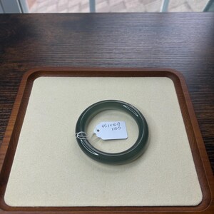 May include: A dark green jade bangle bracelet. The bracelet is circular and smooth, with a polished finish. A small tag with the measurements 55.1 x 10.9 x 10.3 is attached. The bracelet rests on a beige surface within a wooden tray.