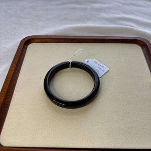May include: A glossy black bangle bracelet, resting on a beige surface within a wooden tray. A small tag is attached to the bracelet. The bracelet's smooth, circular design is highlighted against the neutral background.
