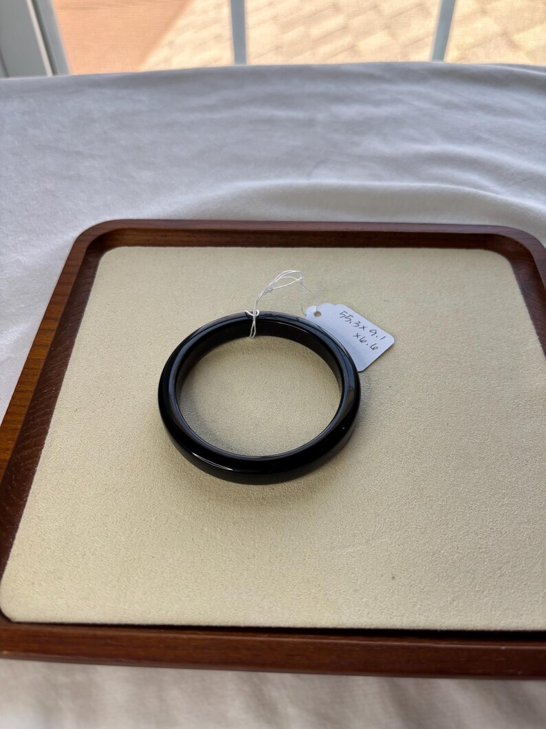 May include: A glossy black bangle bracelet, resting on a beige surface within a wooden tray. A small tag is attached to the bracelet. The bracelet's smooth, circular design is highlighted against the neutral background.