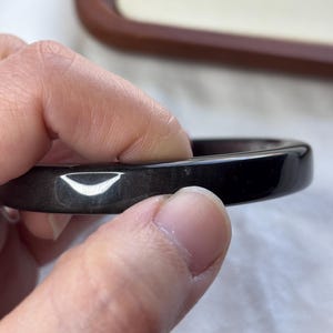 May include: A dark, glossy black bangle bracelet. The smooth, rounded surface reflects light, highlighting its polished finish. The bracelet is held by a hand, showcasing its circular shape and elegant design. A simple, classic piece of jewelry.
