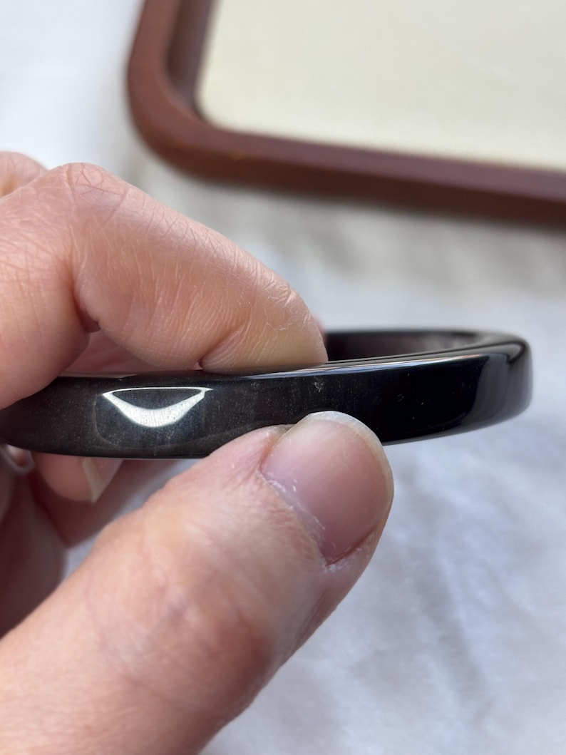 May include: A dark, glossy black bangle bracelet. The smooth, rounded surface reflects light, highlighting its polished finish. The bracelet is held by a hand, showcasing its circular shape and elegant design. A simple, classic piece of jewelry.