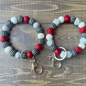 Op de afbeelding: Twee pols-sleutelhangers met kralen. Elke sleutelhanger heeft een mix van rode, witte, grijze en zwarte kralen in verschillende vormen en patronen, waaronder pied-de-poule. De sleutelhangers hebben zilverkleurige hardware.