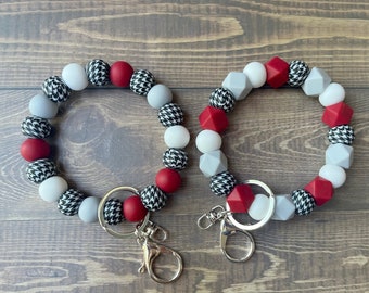 May include: Two beaded wrist keychains. Each keychain features a mix of red, white, grey, and black beads in various shapes and patterns, including houndstooth. The keychains have silver-coloured hardware.