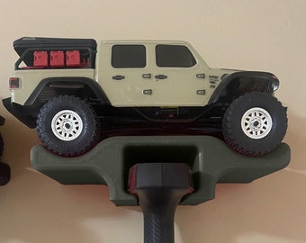 Puede incluir: Un modelo de Jeep teledirigido beige y negro con una baca negra y bidones de combustible rojos. El modelo está montado en un estante verde sobre un mando a distancia negro.